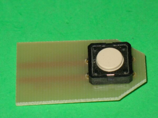 A small white 839-0126 Thumb Button Switch on a green piece of plastic.